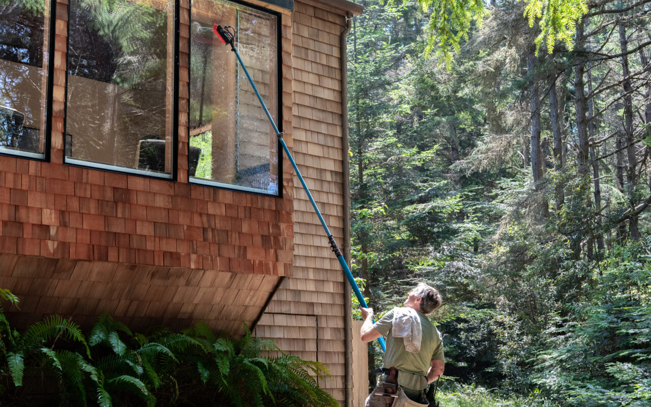 A window cleaner using a water-fed pole on a cedar-shake Northern Minnesota home, surrounded by ferns.